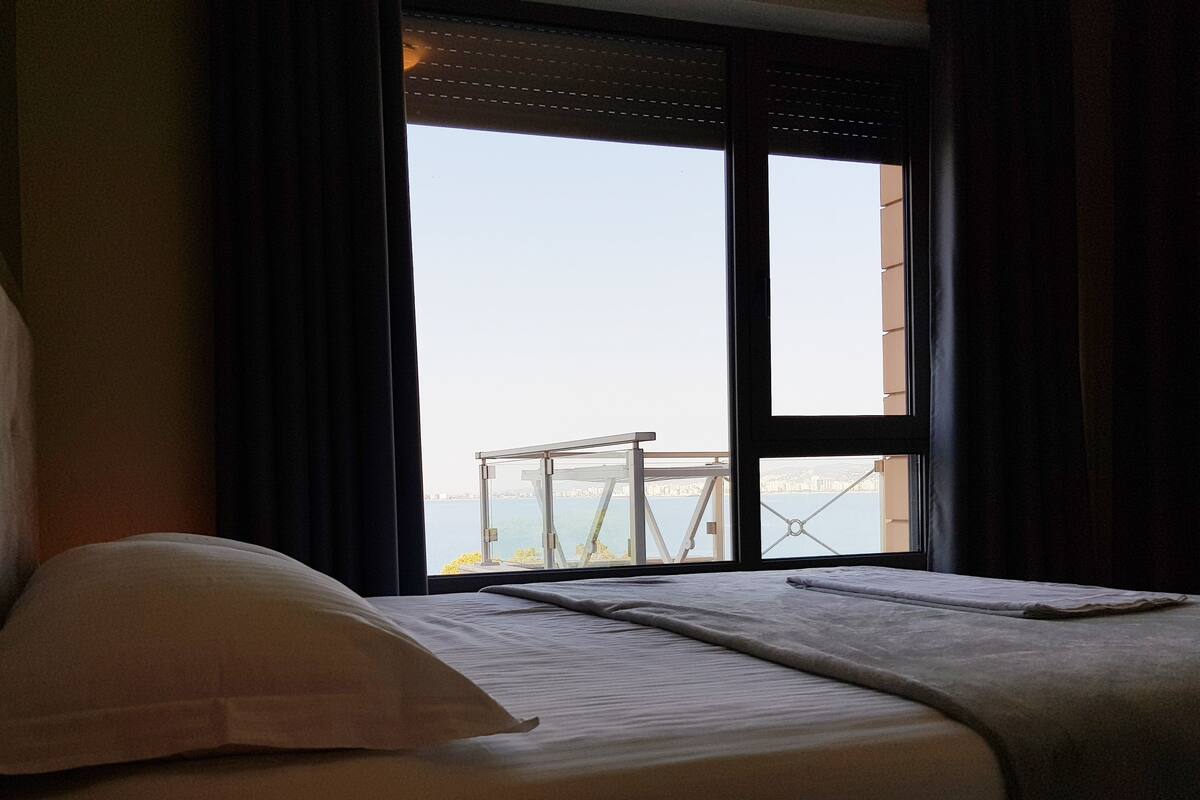 Une chambre d’hôtel avec un lit, une vue sur le paysage urbain à travers une grande fenêtre et un balcon doté d’une balustrade.