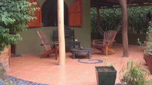 Een overdekt terras met houten meubels, zoals stoelen en een tafel, omgeven door groen en plantenpotten.