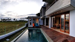 A modern house with a pool, wooden deck, and large glass doors leading to an outdoor seating area. - RC Ubud Villa 1 (Ubud)