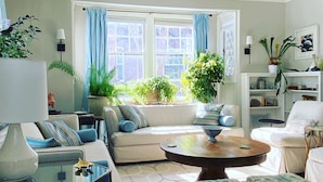 A living room with a sofa, a coffee table, and a patterned ottoman. There are two windows with blue curtains, and a bookshelf with decorative items. - Spacious, Historical, Magical, Designer Home, Prime Walkable Village Location! (Bar Harbor)