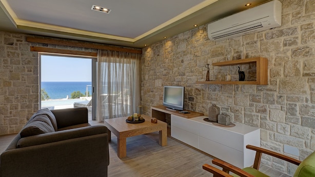 A living room with a stone wall, a television, a wooden coffee table, and a sofa. - Seaside Villa Sfinari – Steps from the Beach (Sfinari)