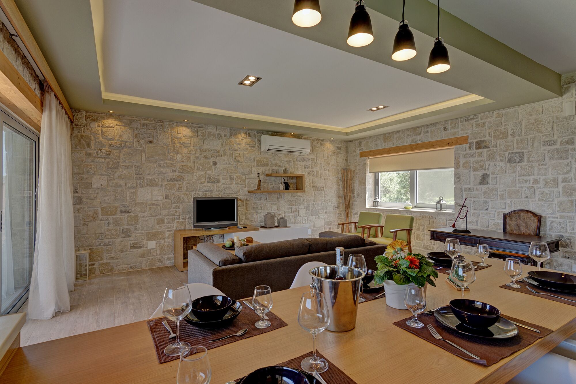 A modern living room with a stone wall, a flat-screen TV, a sofa, and a dining area set for six with wine glasses and a pitcher.
