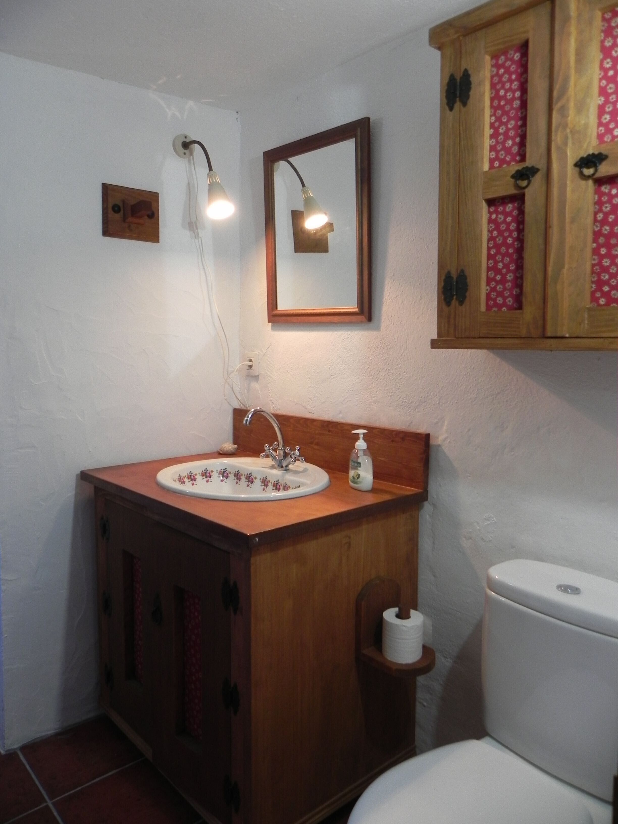 Ein Badezimmer mit einem Holzkosmetikschrank, einem Waschbecken mit Spiegel, einer Toilette und einer wandmontierten Leuchte.