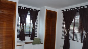 Une chambre à coucher avec un lit, une fenêtre avec des rideaux, une armoire en bois et une étagère avec des vêtements pliés.