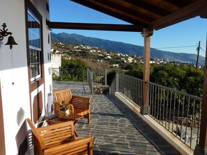 A covered outdoor seating area with stone flooring, wooden furniture, and a view of a hillside town.
