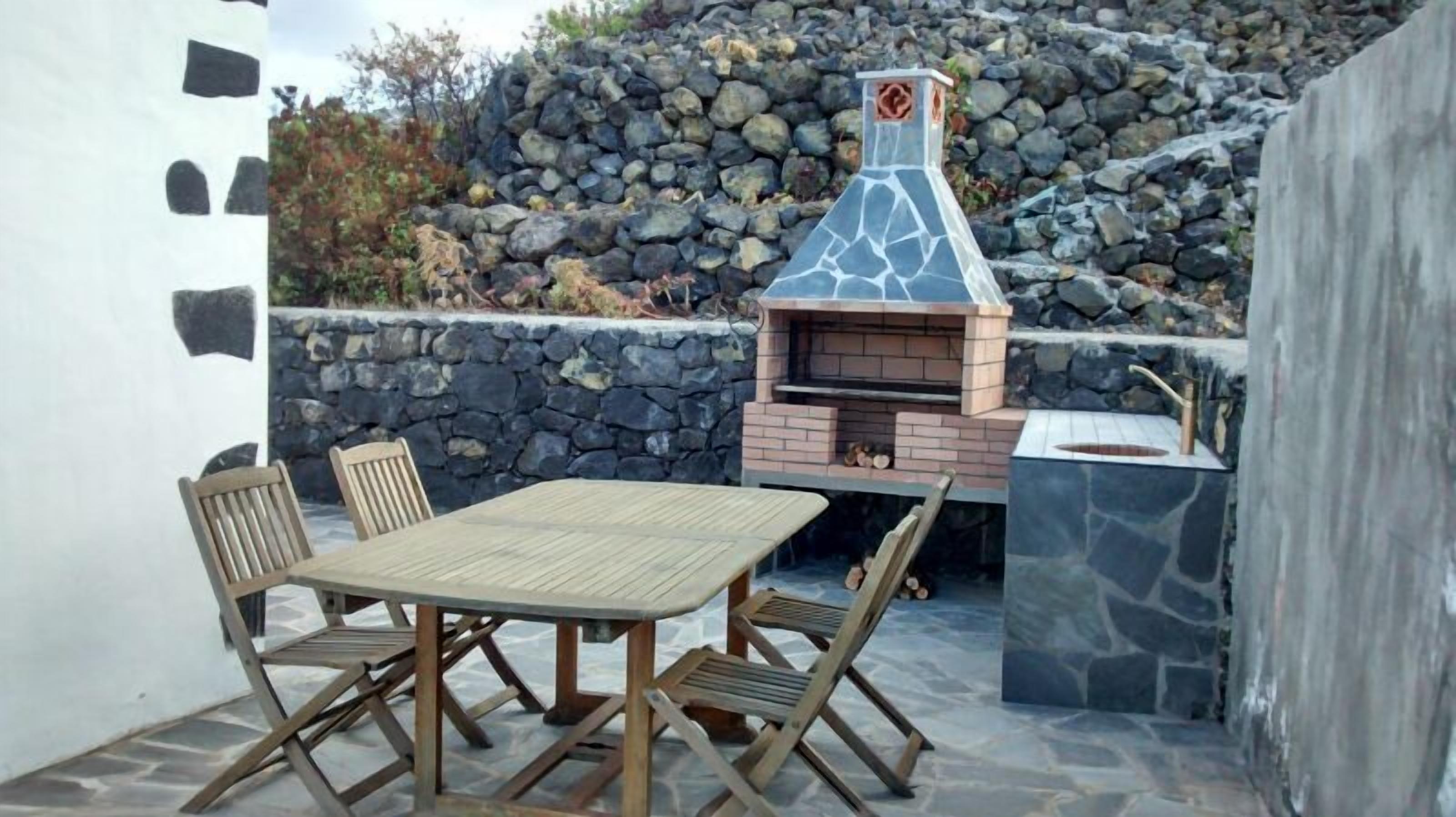 Un patio al aire libre con un muro de piedra, una mesa de madera y cuatro sillas de madera.