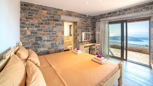 A bedroom with a stone wall, a bed with beige bedding, a desk, a chair, and a view of the sea through a glass door. - Mountain and Private Beach VILLA EAGLE  2-10 pers. (Chavgas)