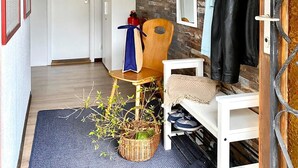 Un pasillo con pared de madera, un perchero con chaquetas, una silla, una cesta con plantas y una puerta que da a otra habitación.