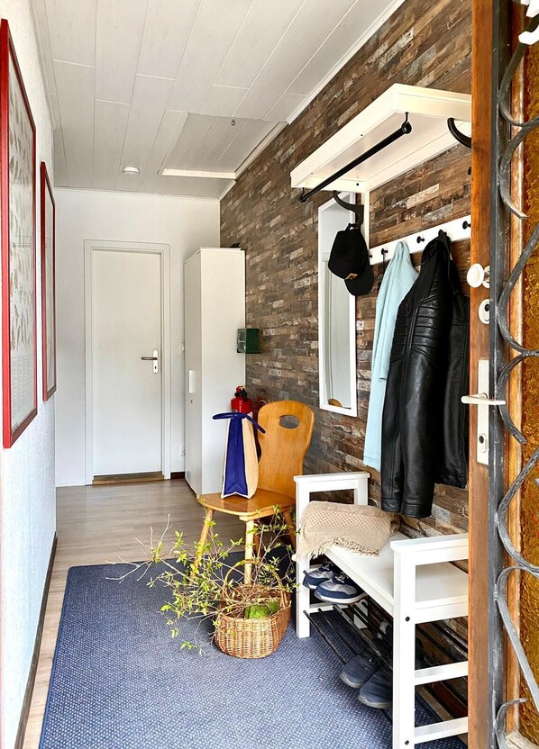 A hallway with a wooden wall, a coat rack with jackets, a chair, a basket with plants, and a door leading to another room.