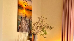 Un dormitorio con cama, almohadas, una mesita de noche con una taza y un libro, un estante en la pared con libros y una planta, y un cuadro sobre el estante.