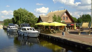 Escena a la orilla del río con barcos amarrados, un edificio con techo de paja y mesas al aire libre bajo sombrillas.