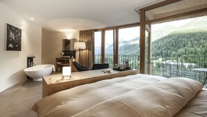 A modern hotel room with a large bed, a wooden desk, a bathtub, and a balcony with a mountain view.