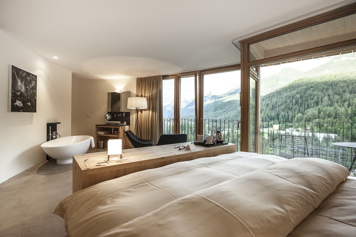Een moderne hotelkamer met een groot bed, een houten bureau, een badkuip en een balkon met uitzicht op de bergen.