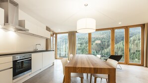 A modern kitchen with a large dining table, ample natural light, and a view of a forested area.
