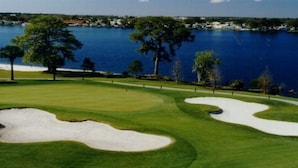 A golf course with sand bunkers, a water body, and a distant shoreline with buildings.