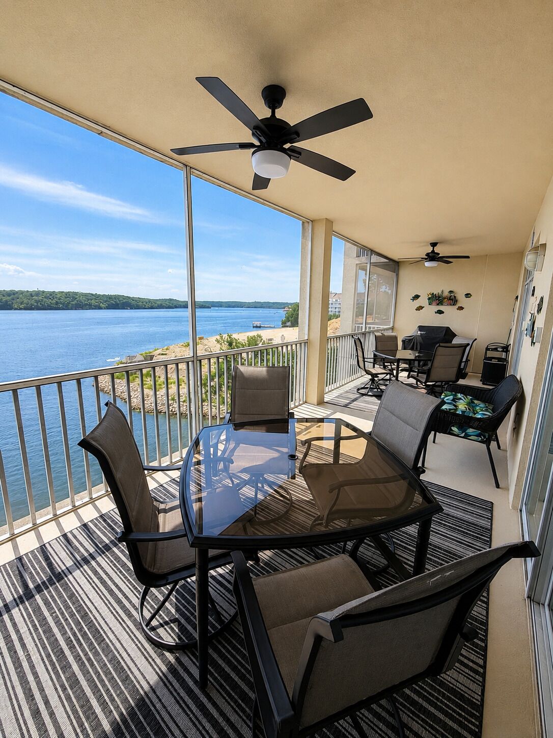 Un balcon avec une table et des chaises en verre, un ventilateur de plafond et une vue sur une étendue d’eau.
