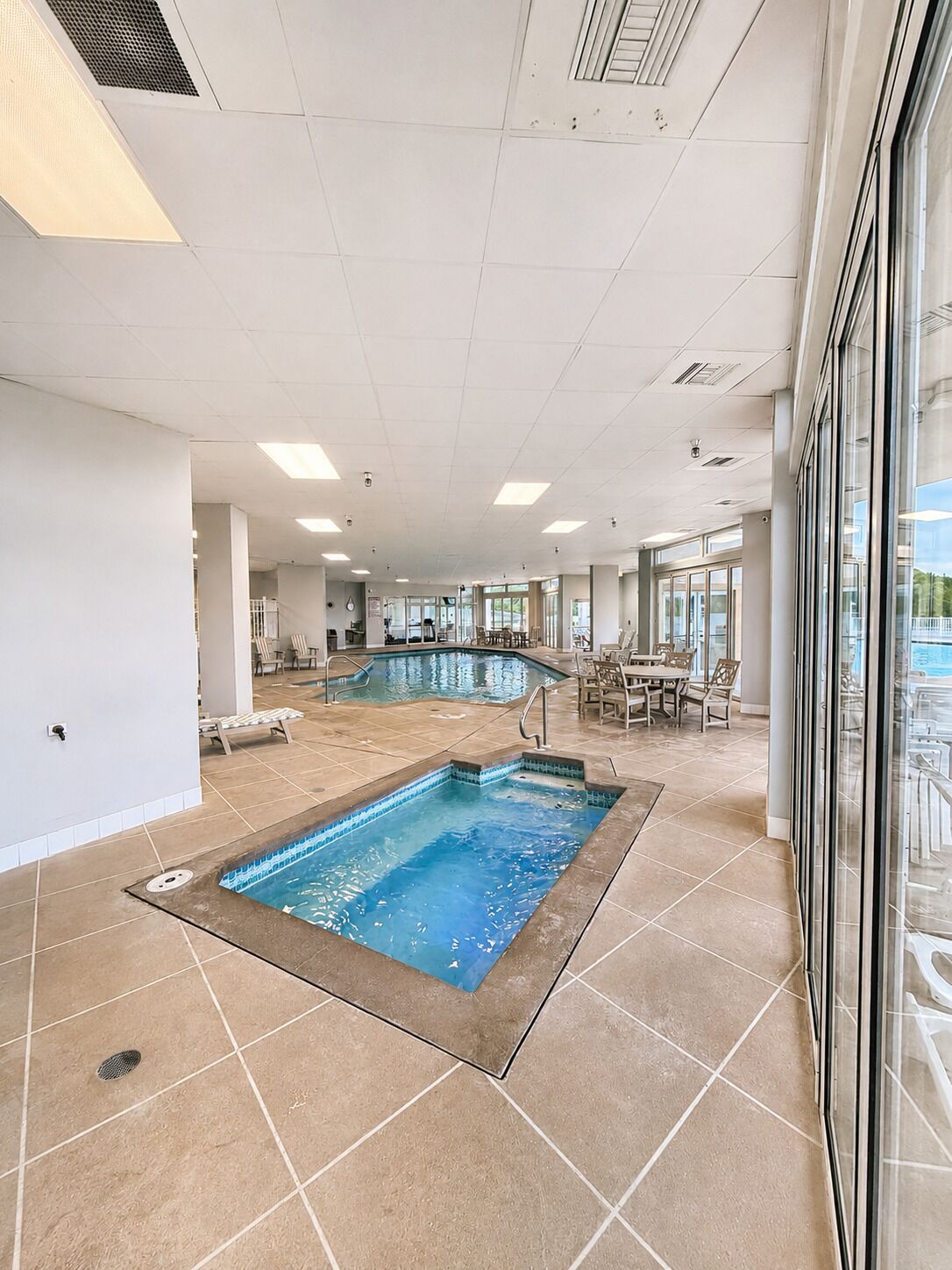 Una piscina con solárium alicatado, zona de estar interior y ventanas amplias.