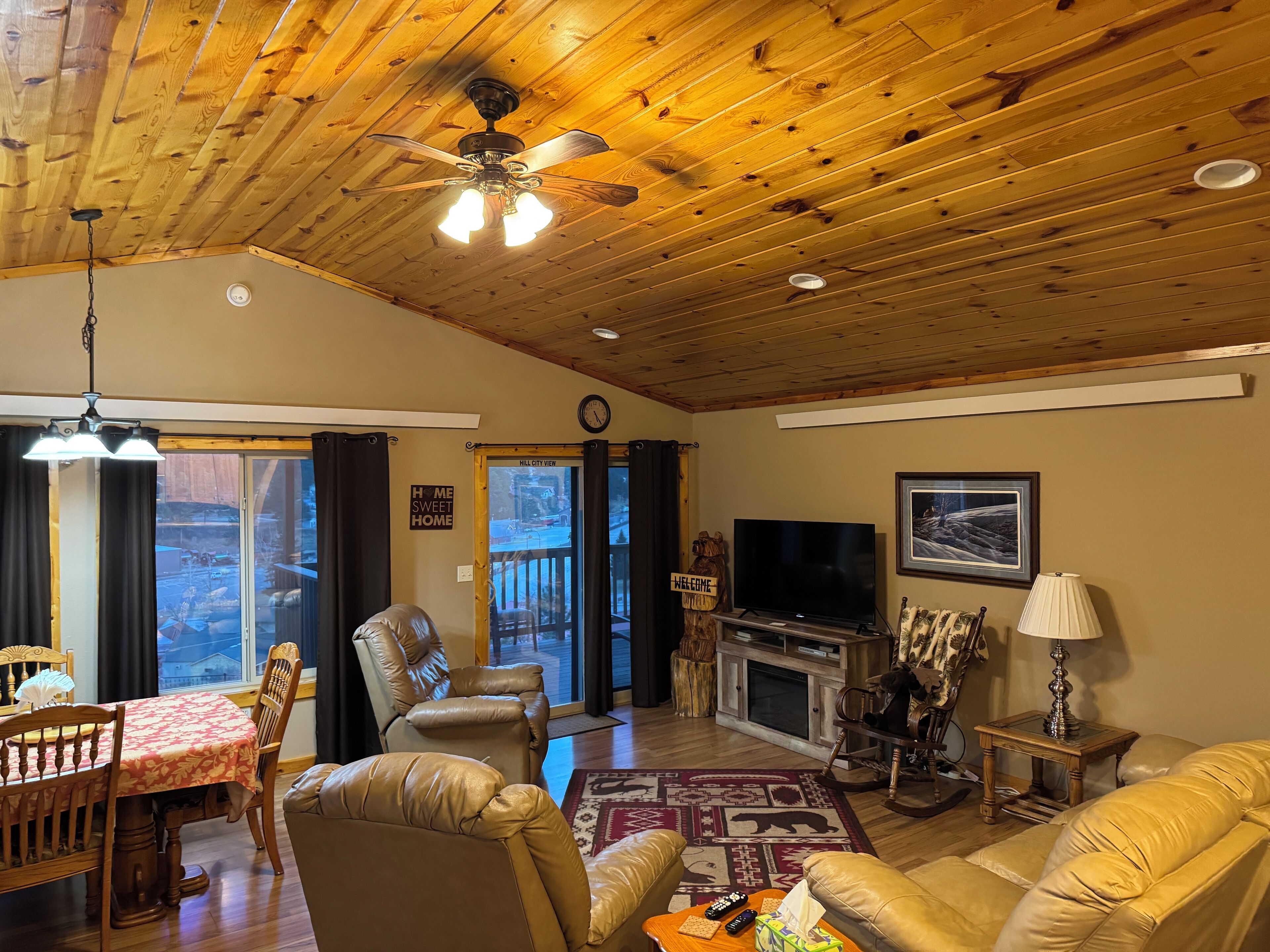 A cozy living room with a wooden ceiling, a television, leather chairs, a sofa, and a fireplace.