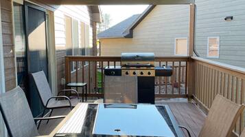 An outdoor patio with a glass table and chairs, a grill, and a view of a house in the background.