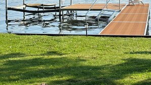 A wooden dock extending into a calm lake with a grassy area in the foreground. - The Bunkalow Lakefront Compound w Lifts for your Speed Boat & Tritoon (Ludington)