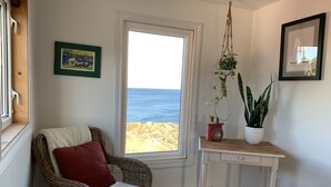 A room with a wicker chair, a small table with potted plants, and a view of the sea through a window. - Oceanfront Cottage at the Edge of the Earth, St. John's  (Pouch Cove)