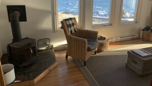 A cozy living room with a large window overlooking the ocean, a fireplace, and a wicker chair. - Oceanfront Cottage at the Edge of the Earth, St. John's  (Pouch Cove)