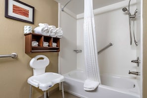 A bathroom with a white bathtub, a shower curtain, a white chair, and a shelf with folded towels. - Comfort Inn and Suites (Alvarado)