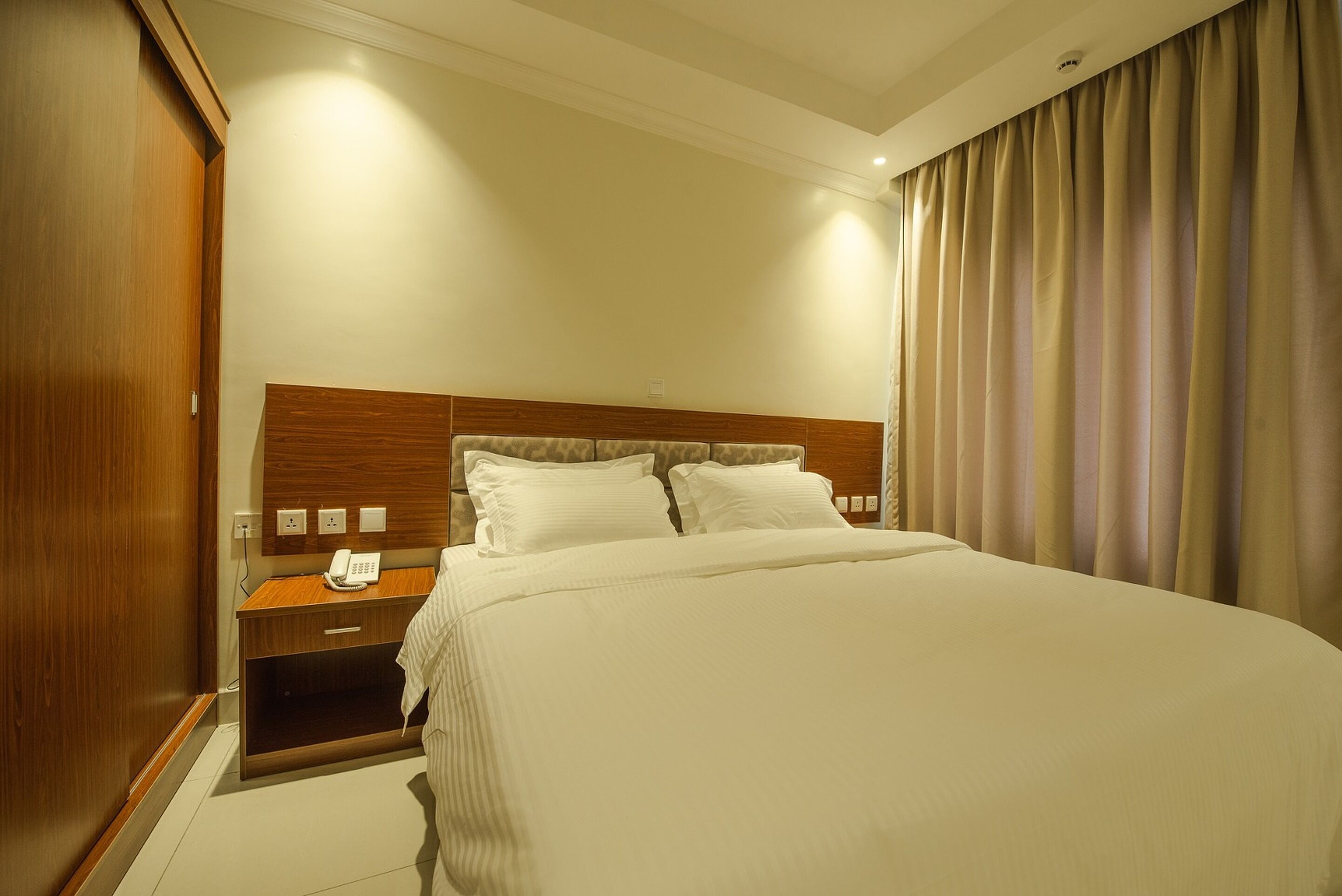 Una habitación de hotel con una cama grande, un cabecero de madera, una mesita de noche con un teléfono y una cortina en la ventana.