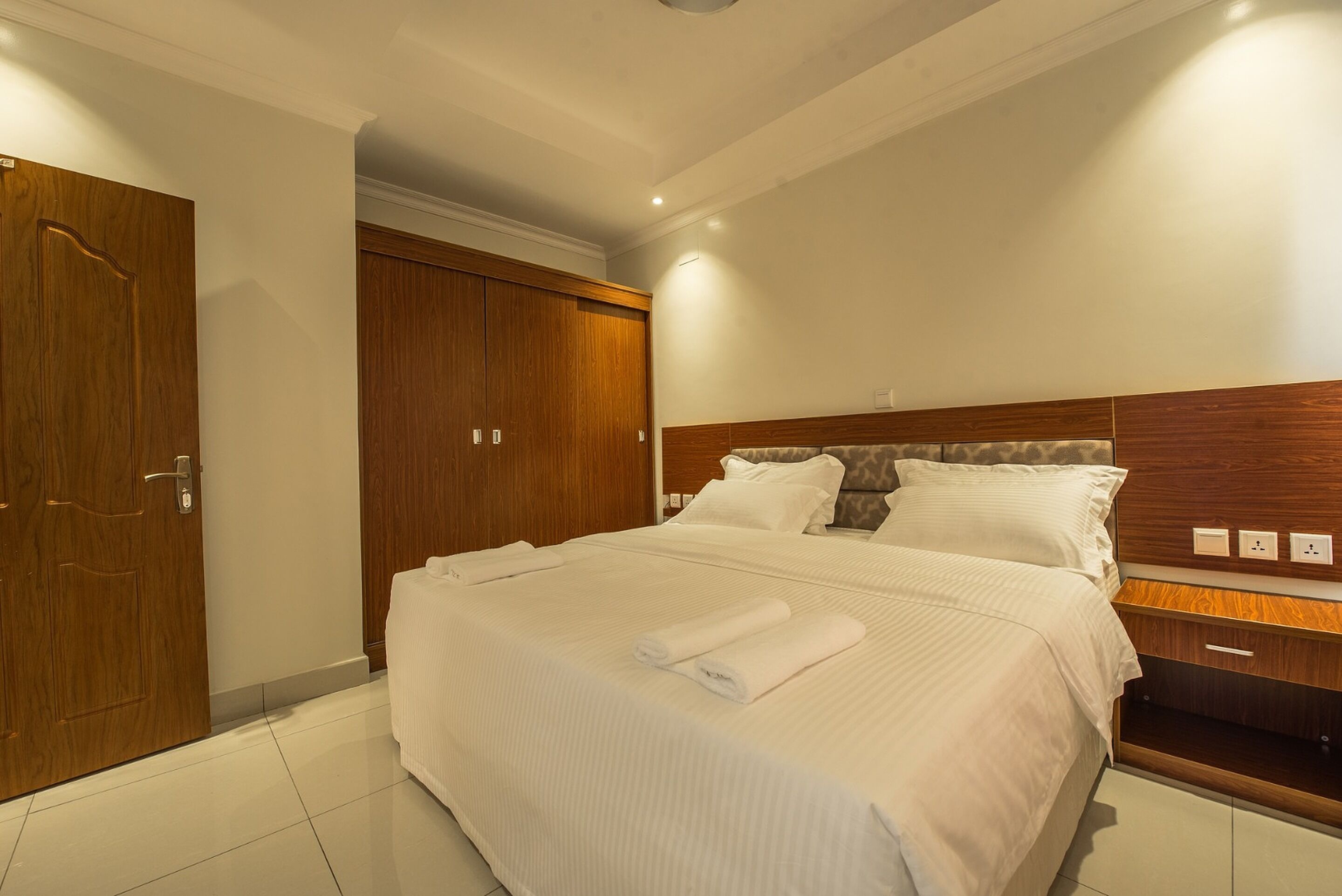 A hotel room with a large bed, wooden wardrobe, and a bedside table with a lamp.