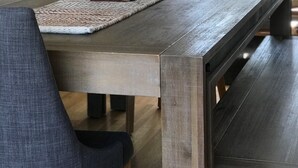 Une table à manger avec un set de table en fibres tressées, deux plantes en pot et une lampe moderne, dans un salon doté d’une grande fenêtre.