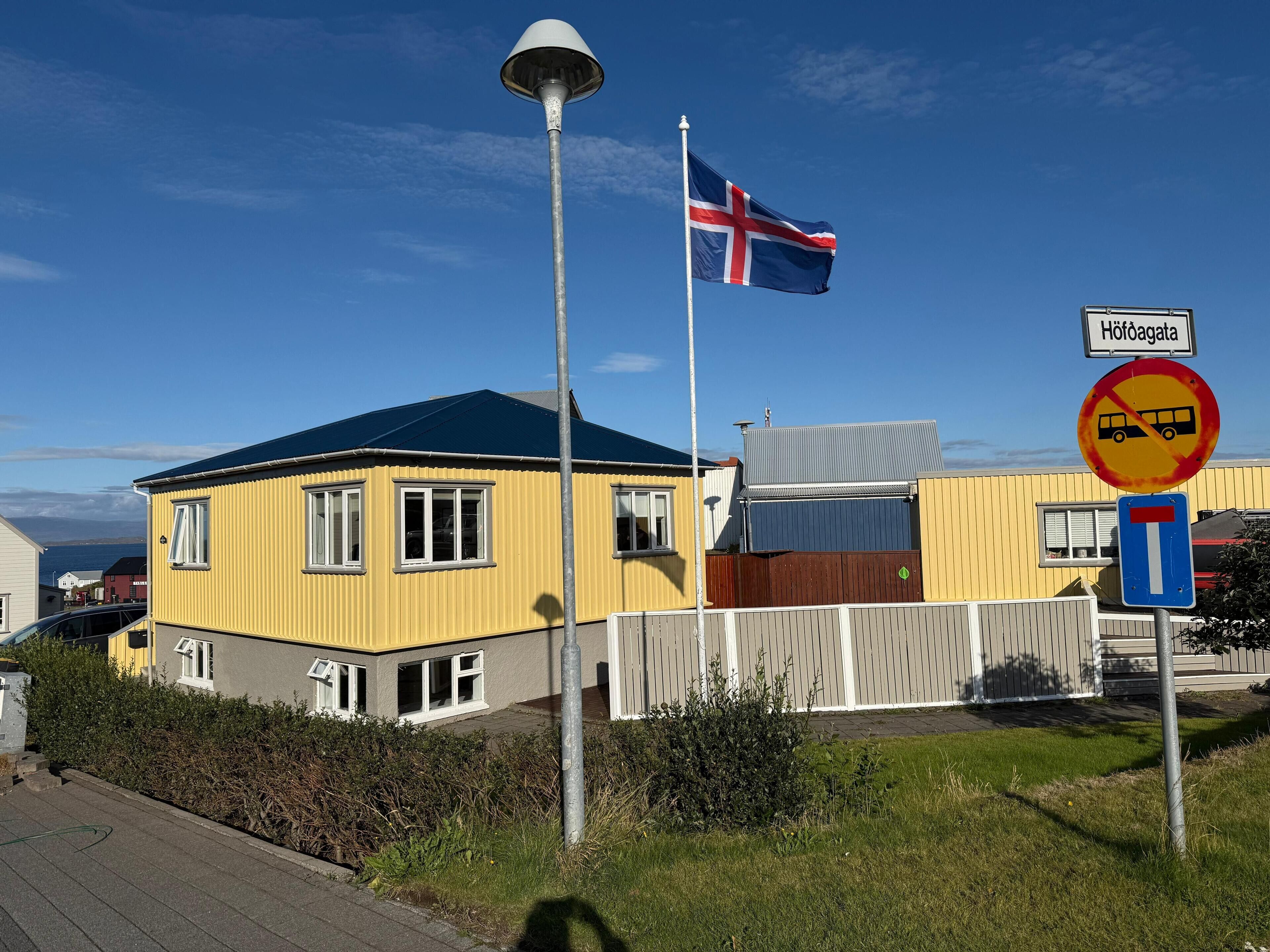 A yellow building with a flagpole, a no bus stop sign, and a street sign.