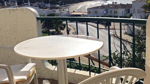 A balcony with a round table and two chairs, overlooking a coastal area with buildings and a beach. - Cozy Apartment with Stunning Sea Views, One Minute Walk from the Beach (Praia da Luz, Lagos)