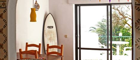 A dining area with a table and chairs, a surfboard leaning against the wall, and a window with colorful bunting.