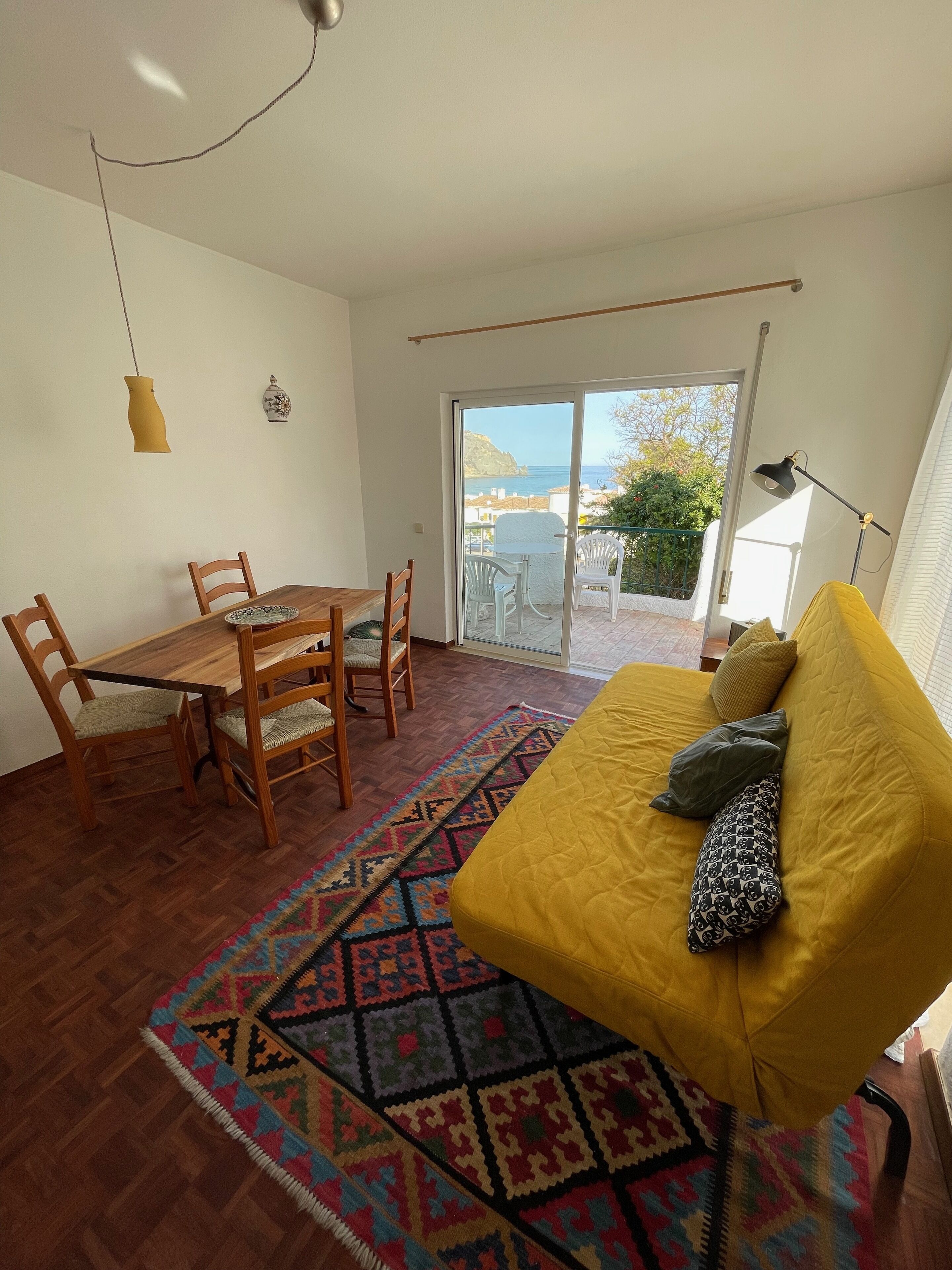 Ein Zimmer mit einem Holztisch, Stühlen, einem gelben Sofa, einem farbenfrohen Teppich und Meerblick durch eine Glastür.