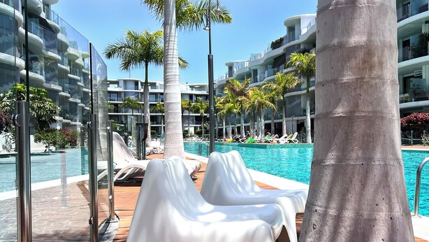 Zona junto a la piscina con sillones de descanso, una palmera y un edificio moderno con balcones.
