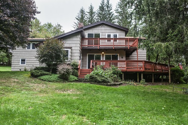 A two-story house with a balcony and a deck, surrounded by trees and a grassy yard.