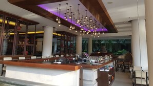 Eine moderne Bar mit einer langen Theke, mehreren hängenden Lampen und Blick auf eine tropische Landschaft durch große Fenster.