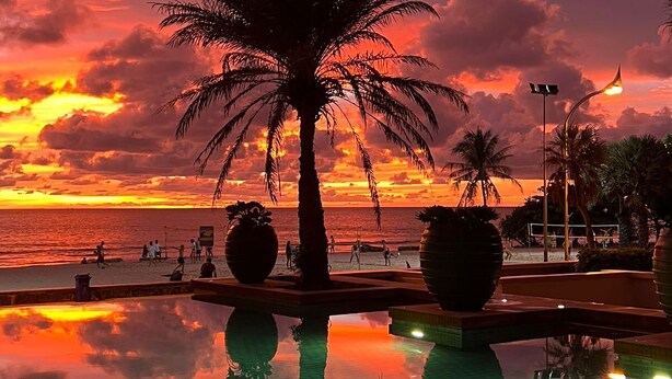 Ein Sonnenuntergang über einem Strand mit Palmen und einem Pool, der den Himmel spiegelt.