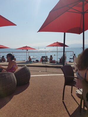 A beachside seating area with red umbrellas and wicker chairs overlooking the sea. - Studio Flat, Sleeps 4. Doorstep skiing with spectacular views and location. (Bernex)
