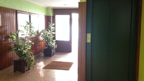 Un couloir aux murs recouverts de lambris en bois, deux plantes en pot, un paillasson marron et une porte verte.