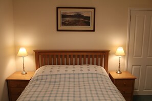 Een slaapkamer met een houten hoofdbord, een geruit beddegoed, twee nachtkastjes met lampen en een ingelijst kunstwerk aan de muur.