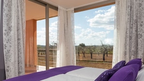A bedroom with a large bed, purple pillows, and a view of an orchard through a sliding glass door.