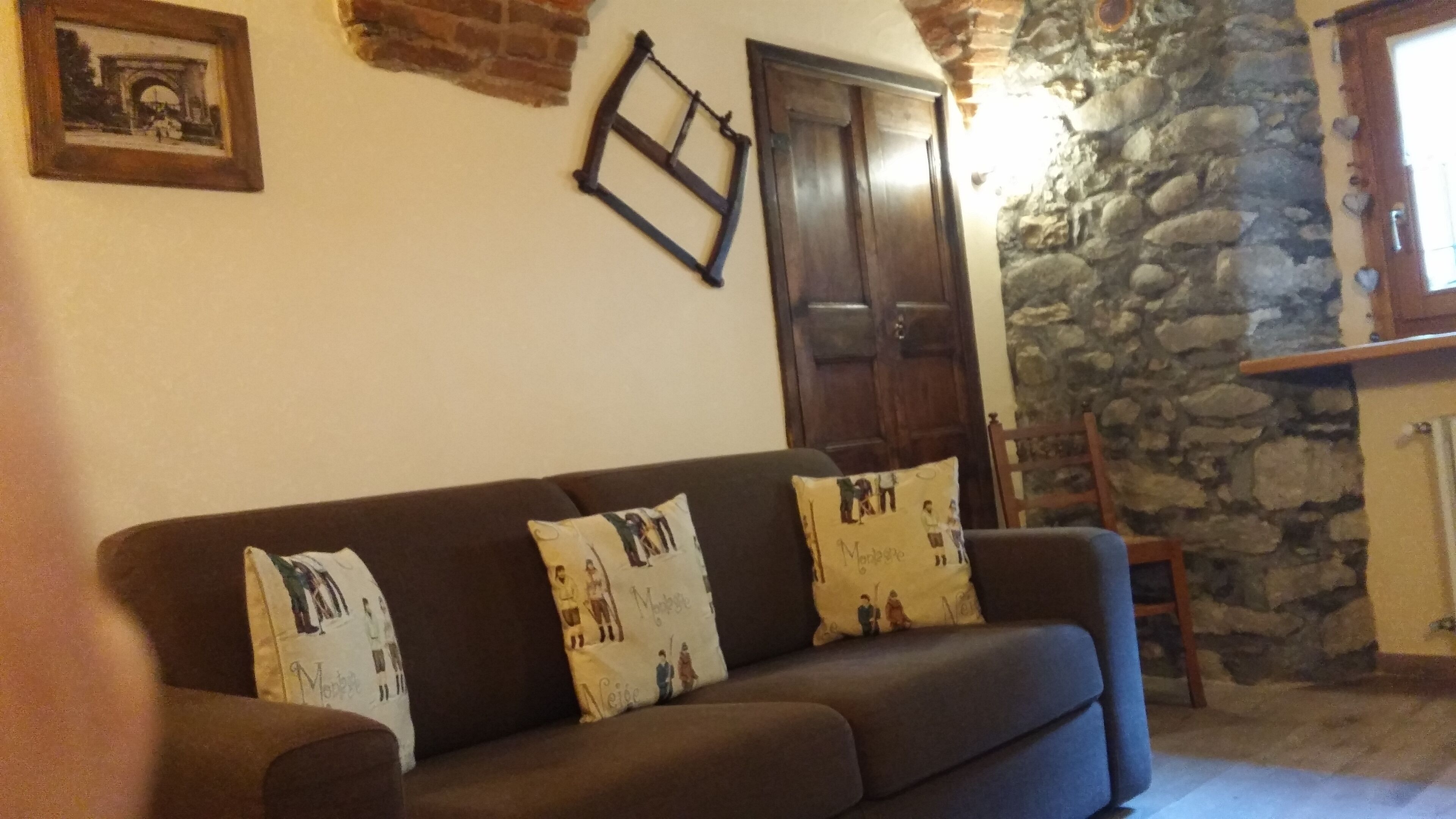 Una sala de estar con un sofá marrón, dos almohadones estampados, una puerta de madera, una pared de piedra y un cuadro enmarcado en la pared.