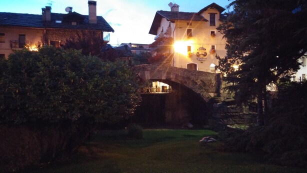 A stone bridge over a small stream, surrounded by greenery and buildings with illuminated signs.