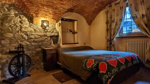 A bedroom with a stone wall, a bed with a heart-patterned blanket, a wooden nightstand, a mirror, and a window with curtains.