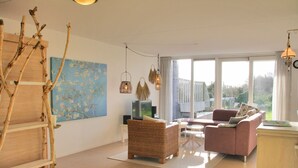 Ein geräumiges Wohnzimmer mit großem Fenster, einer Holzleiter, einem Korbsessel und einem rosa Sessel, sowie einem Flachbildfernseher.