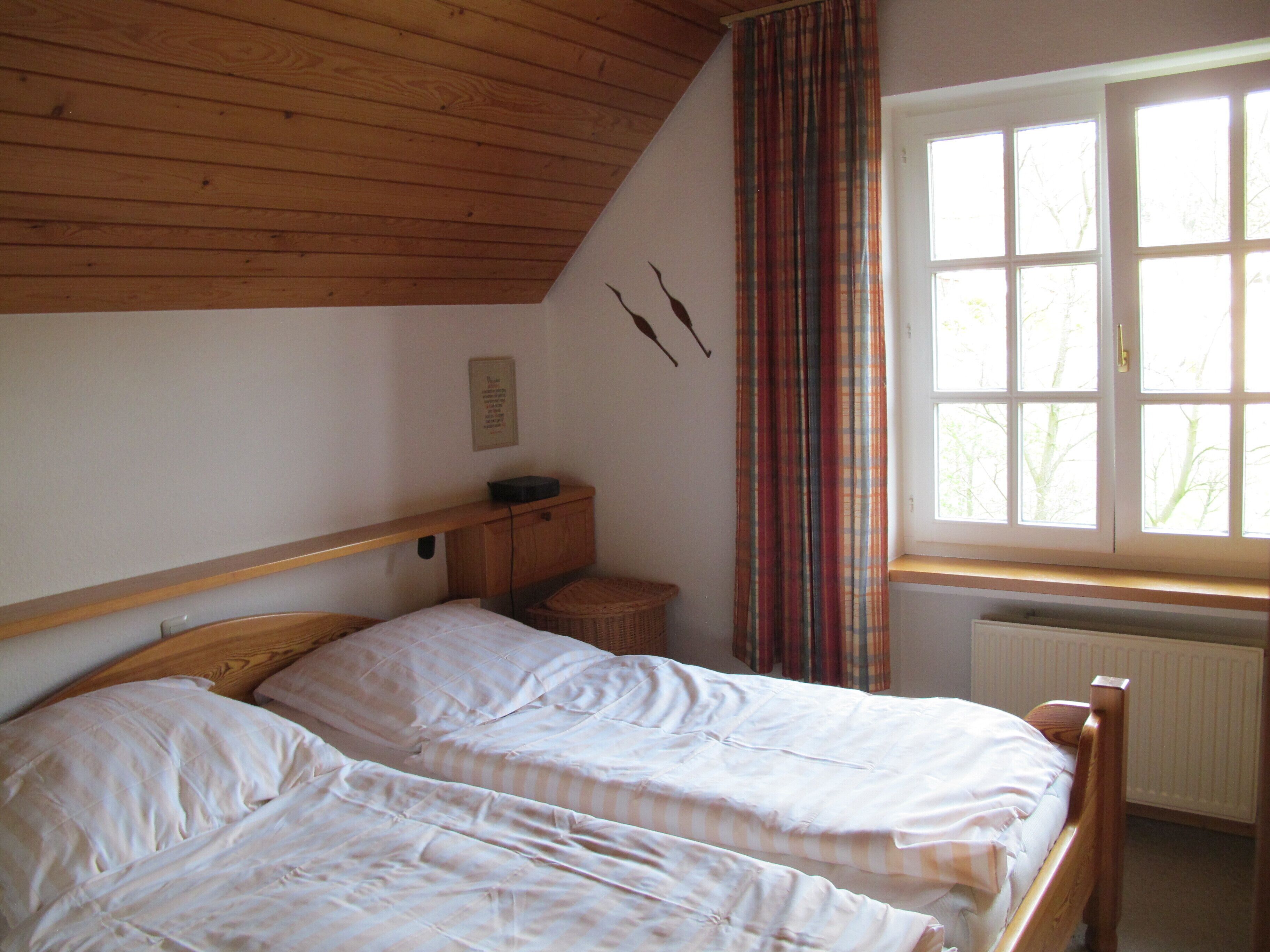 Una camera da letto con due letti, soffitto in legno, una finestra con tende, un termosifone e un piccolo armadietto di legno.