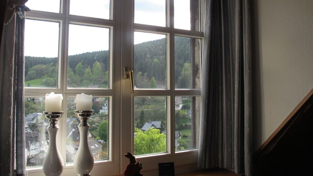 Une pièce dotée d’une grande fenêtre donnant sur une zone boisée et des maisons. Sur une table placée devant la fenêtre, on peut voir deux chandeliers blancs.
