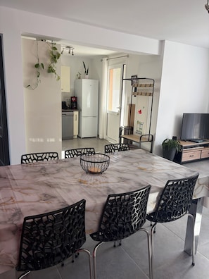 A modern dining area with a marble table, black chairs, a kitchen in the background, and a TV on a wooden cabinet.