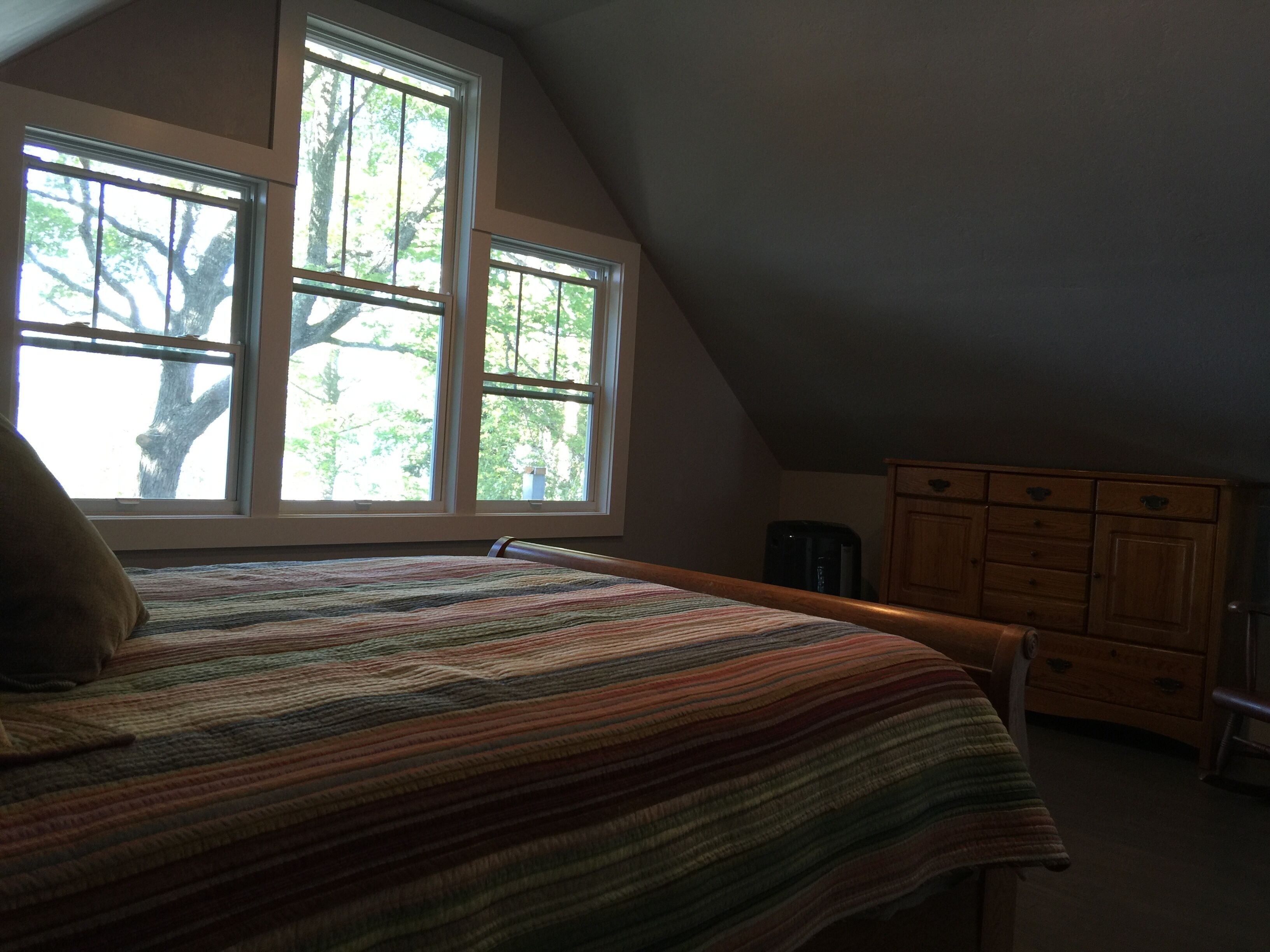 Une chambre à coucher avec un lit, une commode et deux fenêtres donnant sur des arbres.
